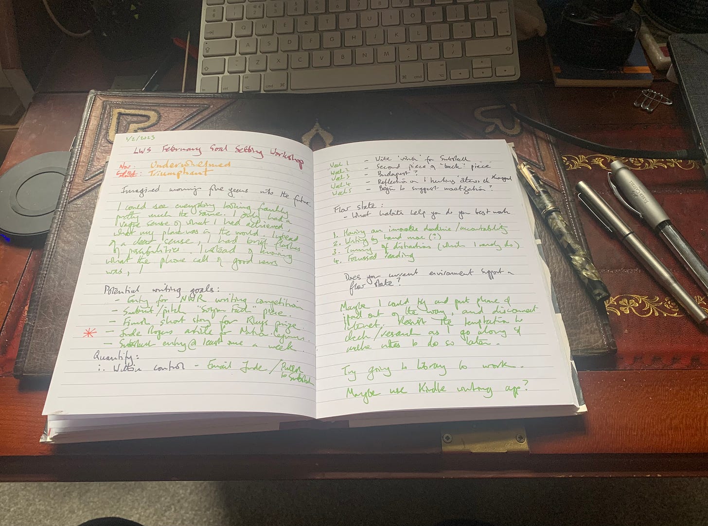 A notebook with handwritten notes in different coloured inks shirts on a desktop with pens and a computer keyboard.