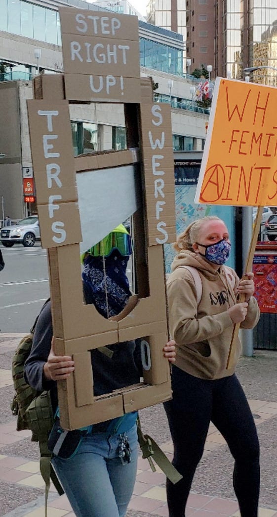 A masked protester outside of the Vancouver Panel on gender identity taunts feminists to "step right up" and stick their head in the guillotine effigy. A masked protester outside of the Vancouver Panel on gender identity taunts feminists to "step right up" and stick their head in the guillotine effigy.