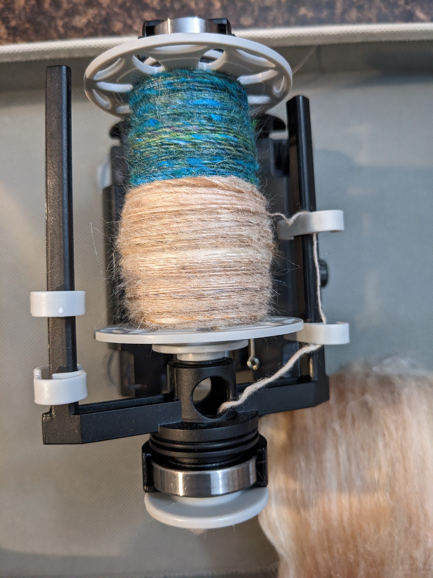 A gray and black electric spinning wheel with two kinds of yarn on the bobbin: one blues and greens and one a light fawn color.
