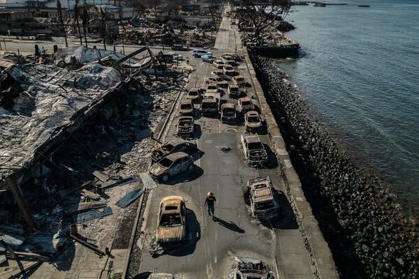 An overhead view shows a police officer walking between wrecked cars on a road along a waterfront. 