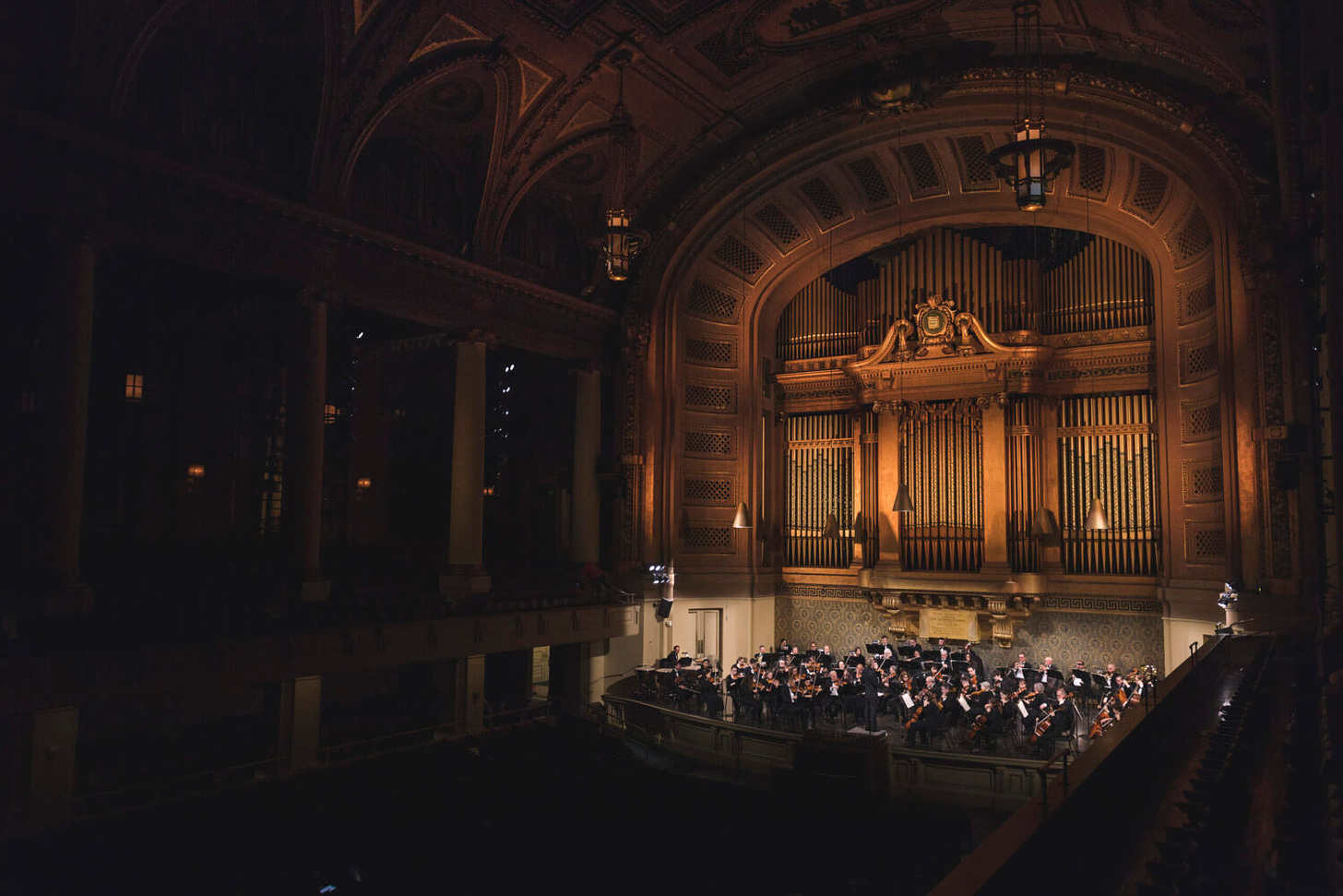 The orchestra plays on the stage at Woolsey Hall. The Hall is in shadows, and the golden organ pipes rise over the orchestra creating a moody and dynamic scene.