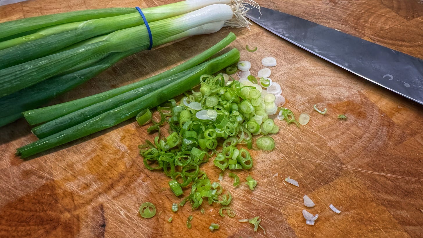 A photo of a bunch of uncut green onions, a pile of sliced green onions, and a chef's knife, all on a cutting board