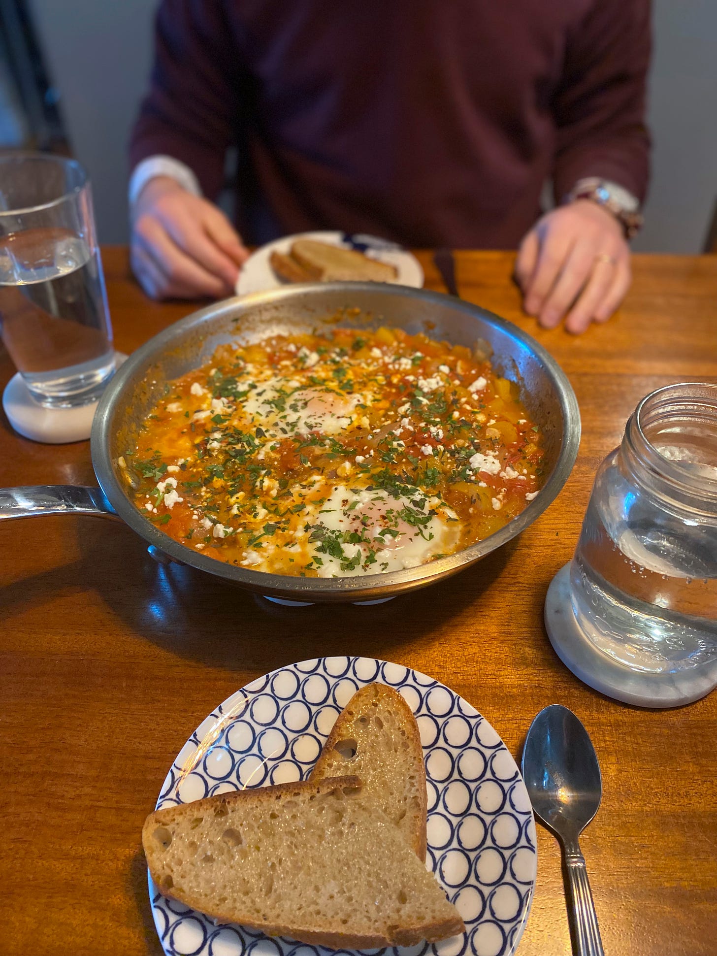 A steel pan of shakshuka, a yellowish-orange colour with splashes of red throughout. Two eggs are poached in the mixture, and the top is sprinkled with feta and a mix of herbs. On opposite sides of the pan are side plates with slices of bread. Jeff sits in front of the plate in the background, his hands resting on the table.