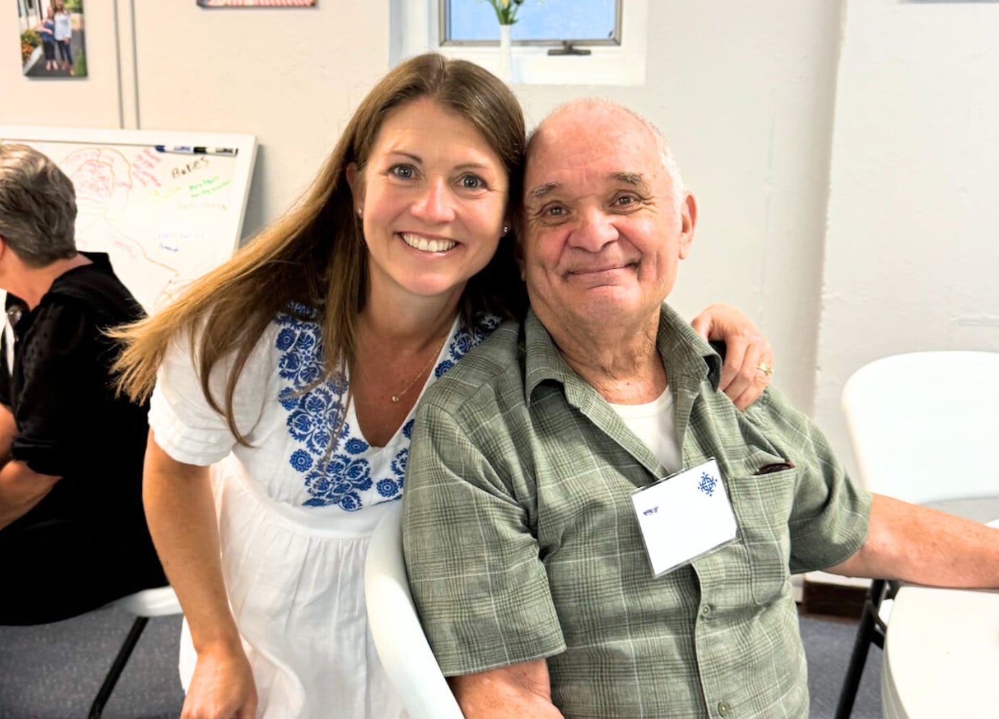 Amy Julia bends down and puts her arm around an older man who is sitting a table for a potluck. Their heads are close together and they are smiling.