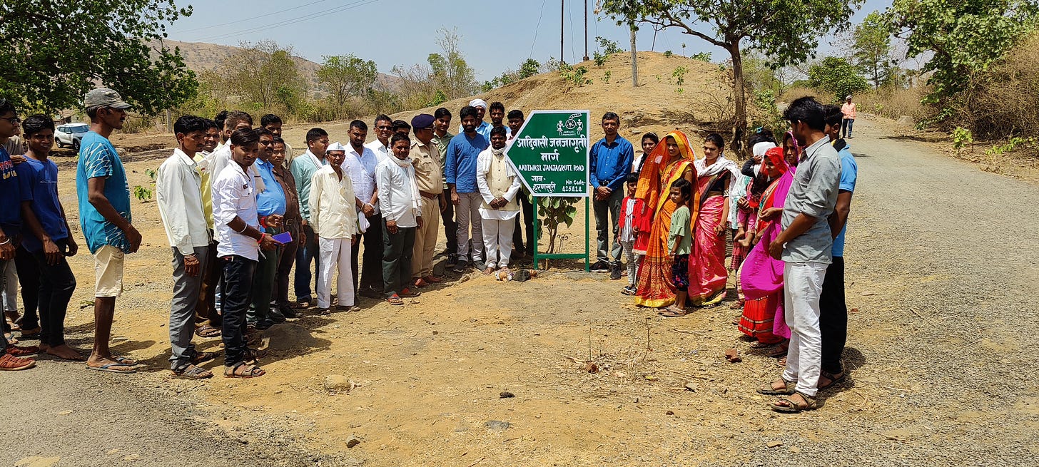 A group of people comprising of the Aadiwasi Janjagruti team and their volunteers standing around a plaque on a road named after their organisation