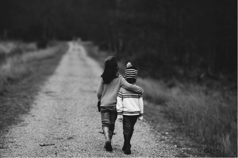 Black and white image of two children seen from the back, arms around each other, walking down a country road.