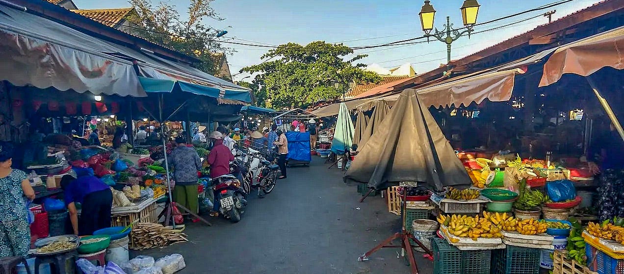 Stalls set up across from each other filled with produce on one side, dry goods on the other. People walk or drive around on their mopeds. 