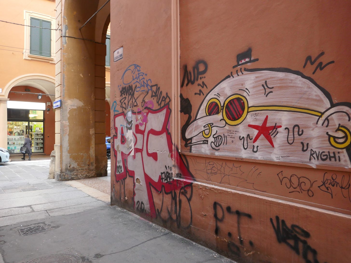 An orange wall with street art including a creepy-looking round-headed man, with a pedestrian entering a shop in the background