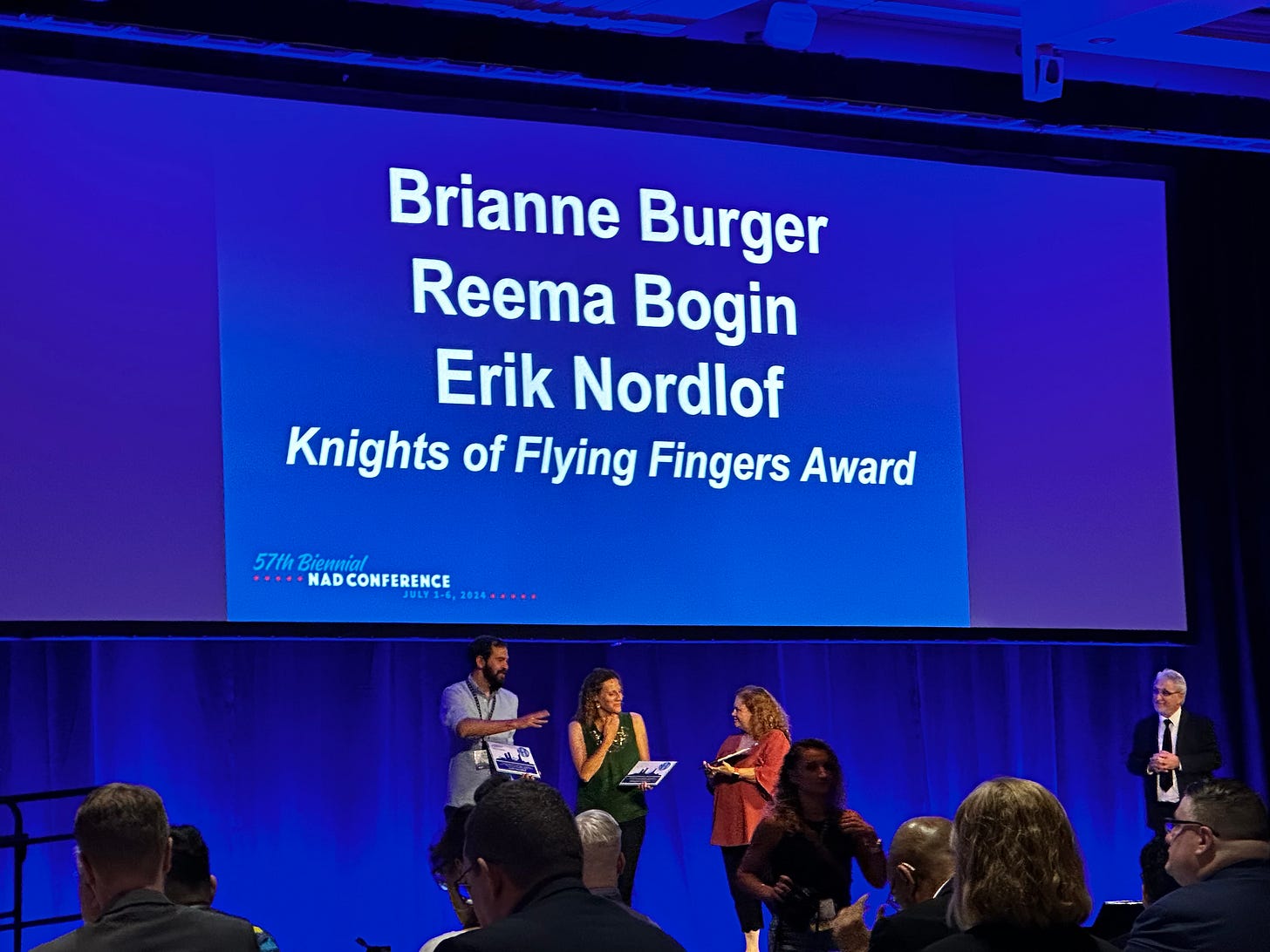 Erik Nordlof, Reema Bogin, and Brianne Burger stand in front of a bright blue projector screen at the NAD conference, receiving their awards. Their names are listed on the screen, as well as "Knights of Flying Fingers Award."