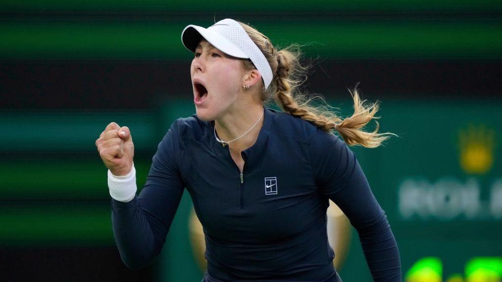 Mirra Andreeva wearing a navy long-sleeve top, white Nike visor, and a braided ponytail clenches her fist and shouts in celebration. Her face shows intense emotion, and the blurred green background features a Rolex logo, indicating a professional tournament setting.