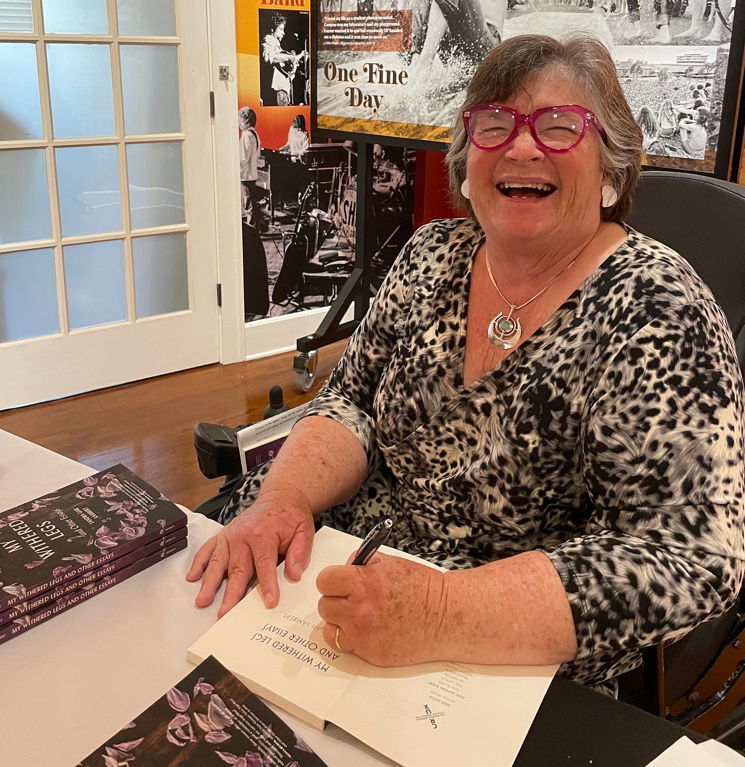 An older white woman smiling big and signing books. She's wearing a black and white dress, a silver necklace, white enamel earrings, and pink glasses. 