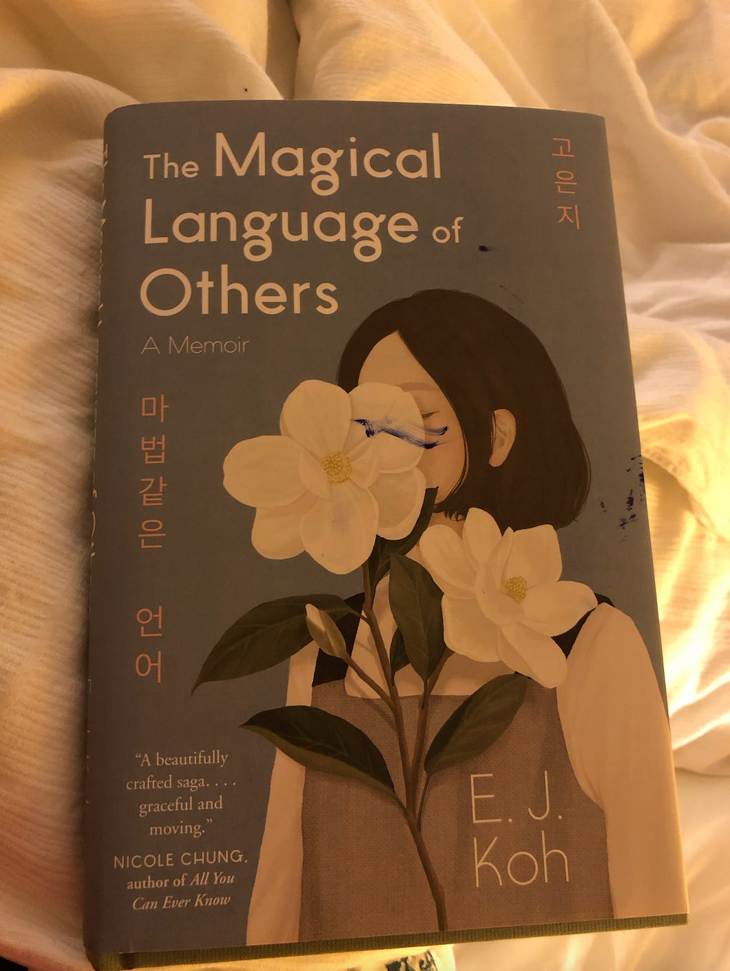 A photo of Kat’s copy of The Magical Language of Others by E.J. Koh. The cover has a drawing of a woman whose face is obscured by a beautiful white flower. The text has the title and author’s name written in both English and Korean.