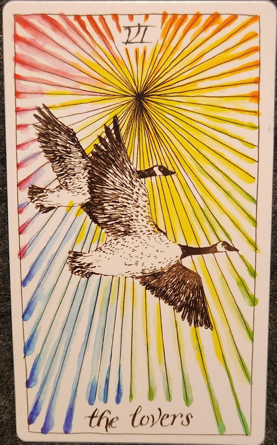 A tarot card labelled VI the lovers. Two Canada Geese are flying across the cards from left to right. Their wings spread out in identical formation. The background of the card is a rainbow of colourful lines radiating from a black focal point. 