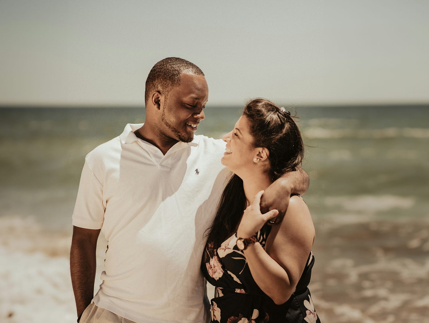 A man and woman smile at each other on a sunny beach. The man has his arm around the woman