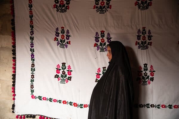 A woman in a dark robe and head covering stands in profile against a white sheet embroidered with stylized flowers.