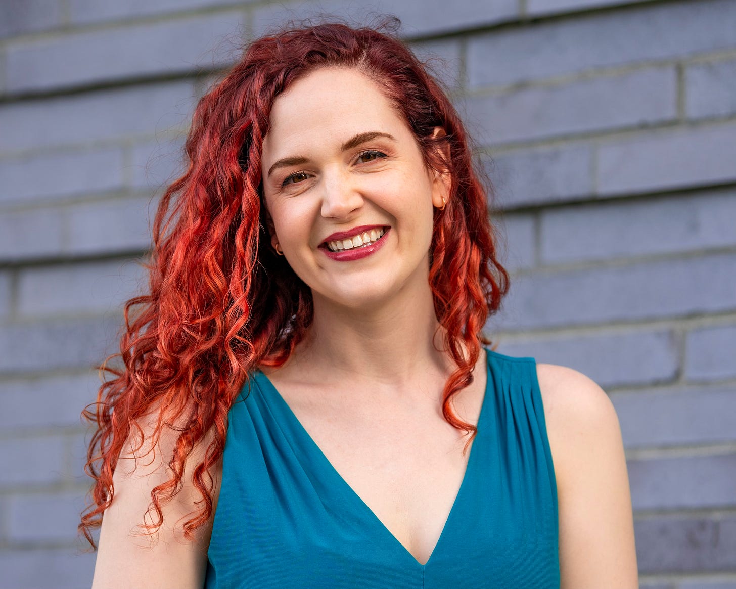 A woman with red curly hair and a teal dress stands in front of a brick wall, smiling. 