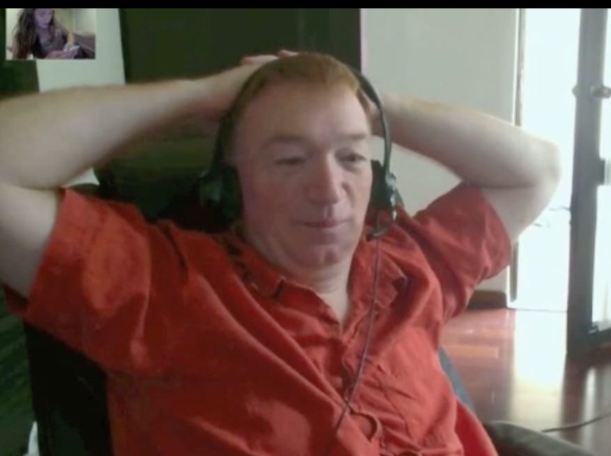 Still from a screen recording of a skype call. In the upper left window, Louise is lying on a bed looking at her phone. Most of the frame is occupied by Scott, who is leaning back in his swivel desk chair holding his hands behind his head and smiling. 