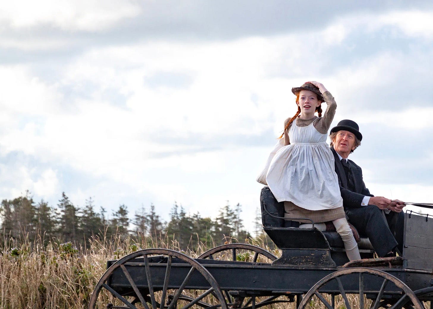 Man and child in 1890s attire riding a carriage through a field. The child looks in the distance, holding her hat.