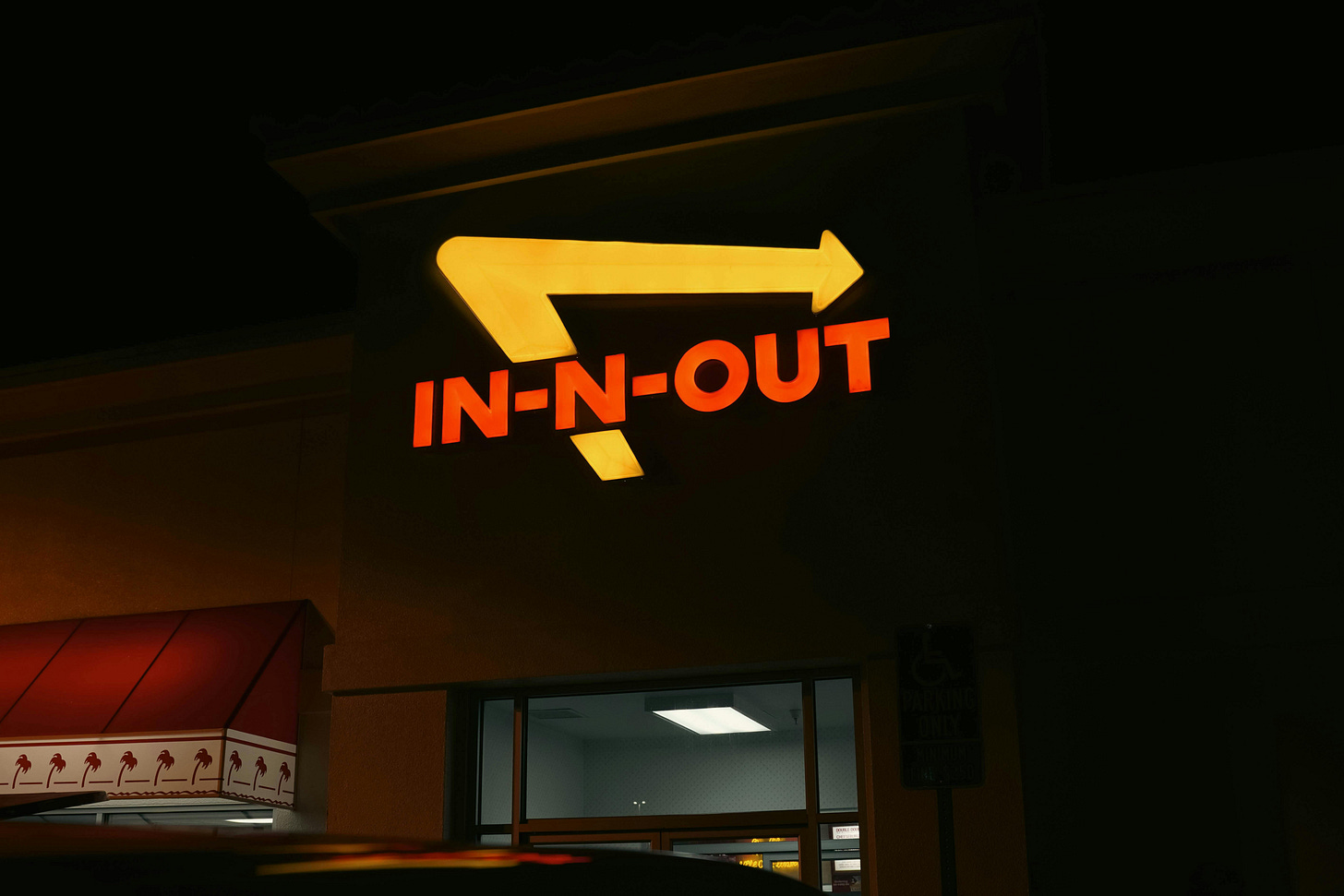 This image shows the iconic In-N-Out Burger restaurant sign illuminated at night. The bright yellow arrow with the red &quot;IN-N-OUT&quot; text stands out against the dark background of the restaurant exterior. A portion of the restaurant&#39;s trademark red awning with palm tree designs is visible in the lower left corner, and the store&#39;s illuminated interior can be glimpsed through the windows below the sign.