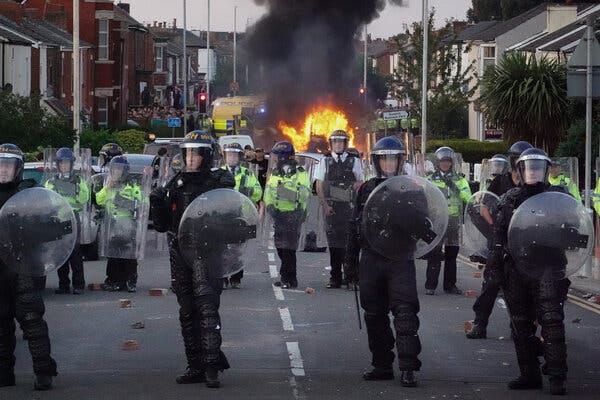 A group of police officers in riot gear, holding shields, on a residential street. A car is on fire behind them.