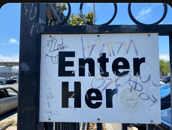 Image of a street sign that reads originally "enter here" but some wag has altered it to white out the second e so it reads 'enter her" coated with some tags in black marker, on a black metal fence, cars and sky are viewable behind.