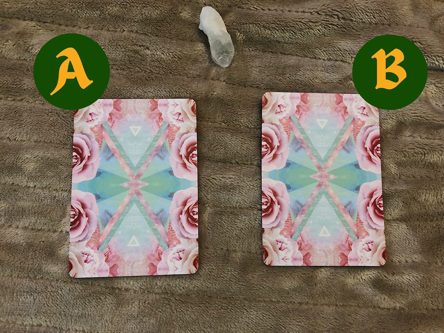 Two pastel colored oracle cards face down with an "A" on the left card, and "B" on the card on the right. Both cards are sitting on a heated beige blanket with a clear crystal between them both