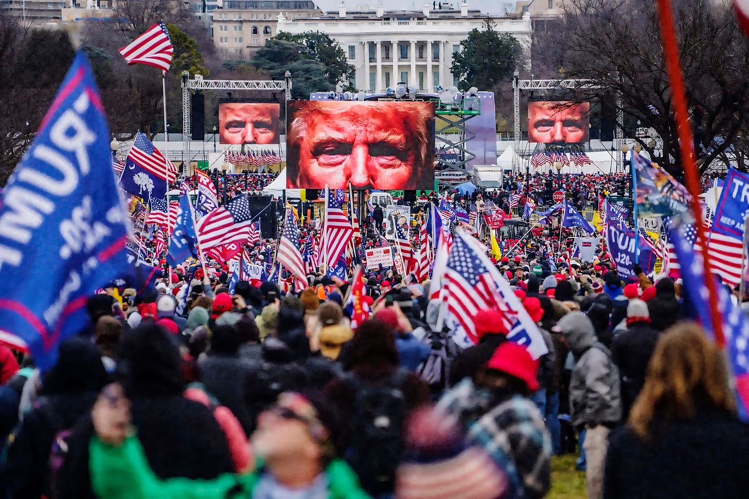 A large crowd of Trump supporters at a rally in Washington, D.C., waving American flags. Two large screens display an image of Donald Trump's face in the background