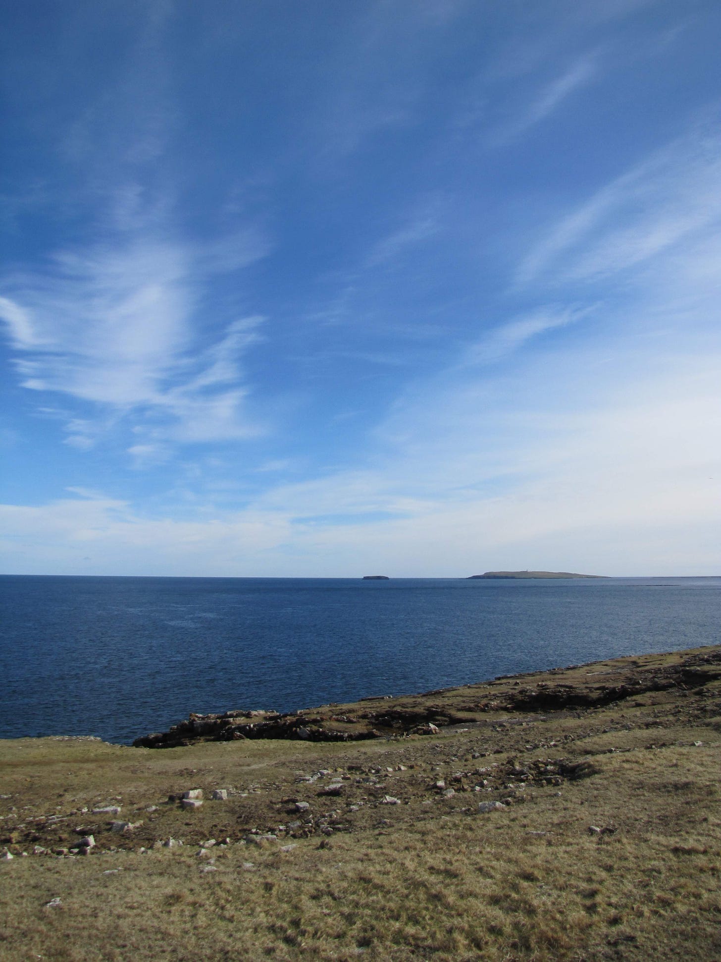 Copinsay and the horse of Copinsay in Orkney. There are two islands in the sea, with a large blue sky above, and the clifftops of Deerness in the foreground.