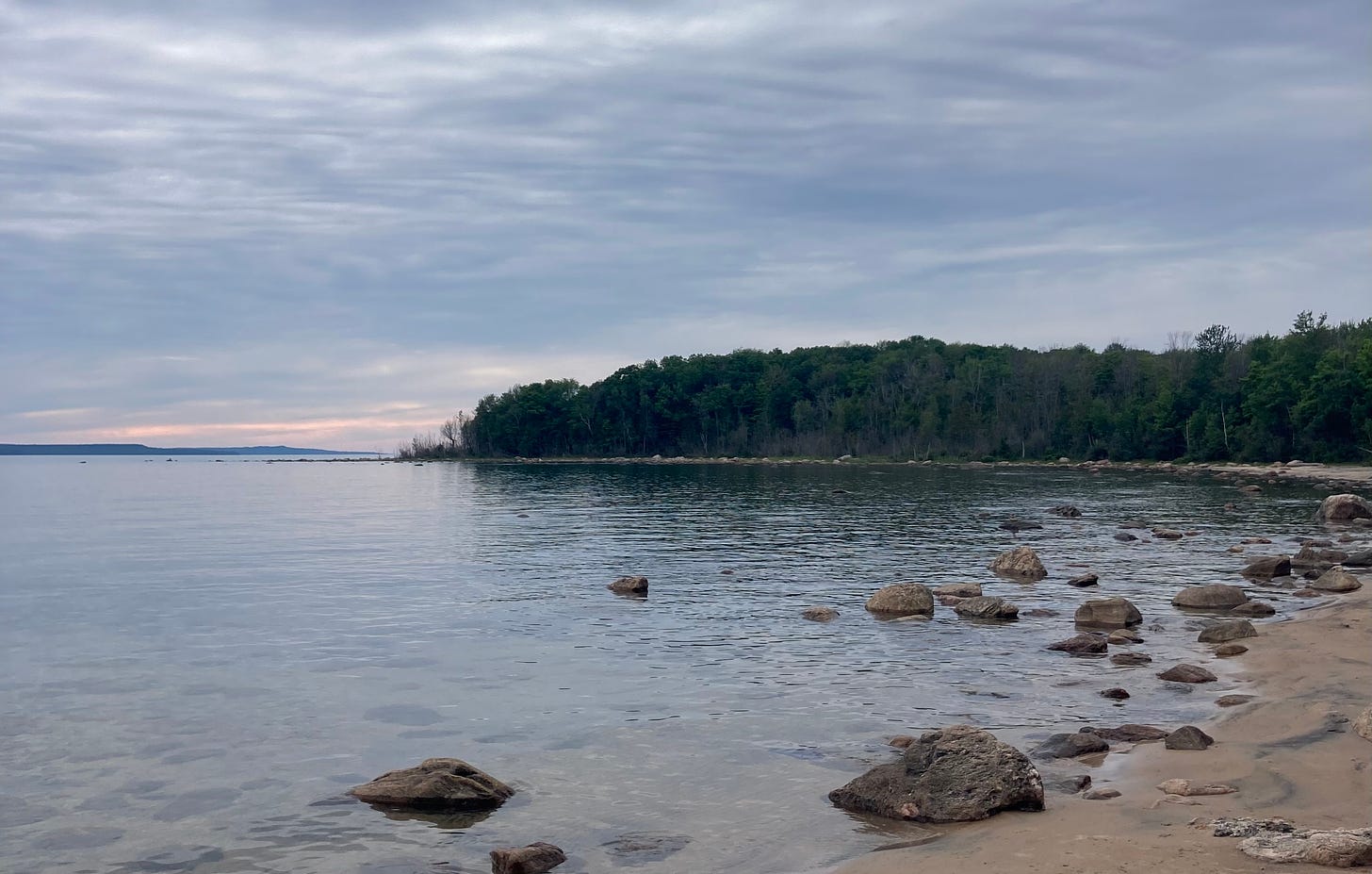 A sandy beach with many large rocks in the water. A treed peninsula stretches into the water. The sky is cloudy with the hint of a sunset