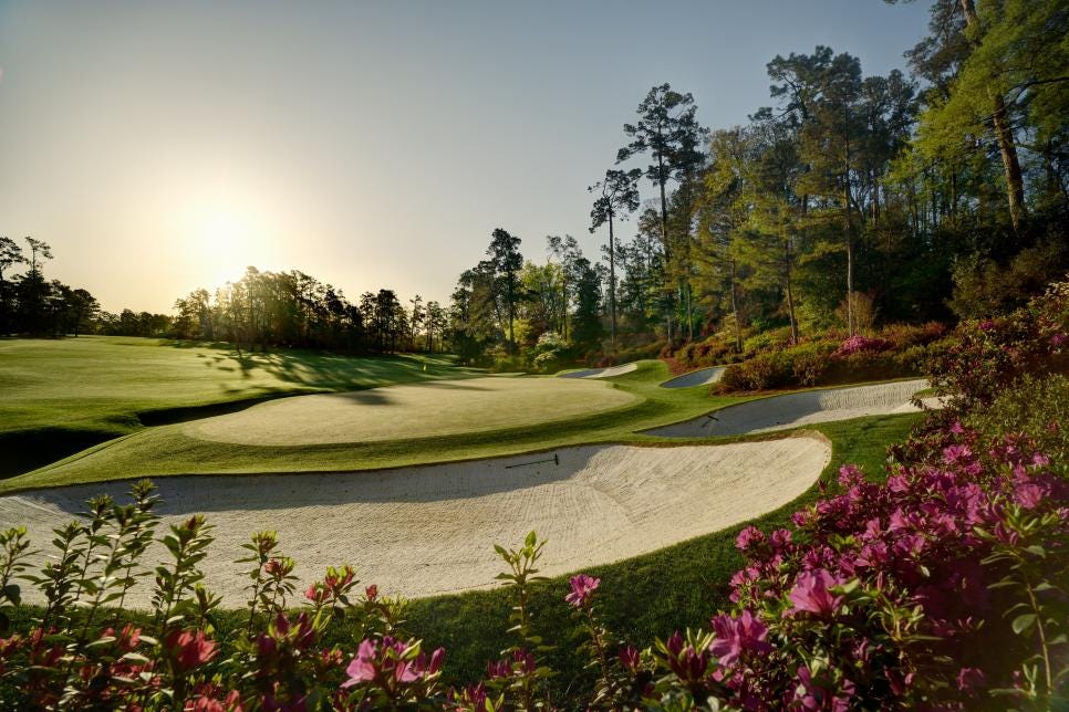 A beautifully manicured golf hole with white sand bunkers, azalea flowers in bloom, and tall pine trees lining the fairway, bathed in golden sunlight.