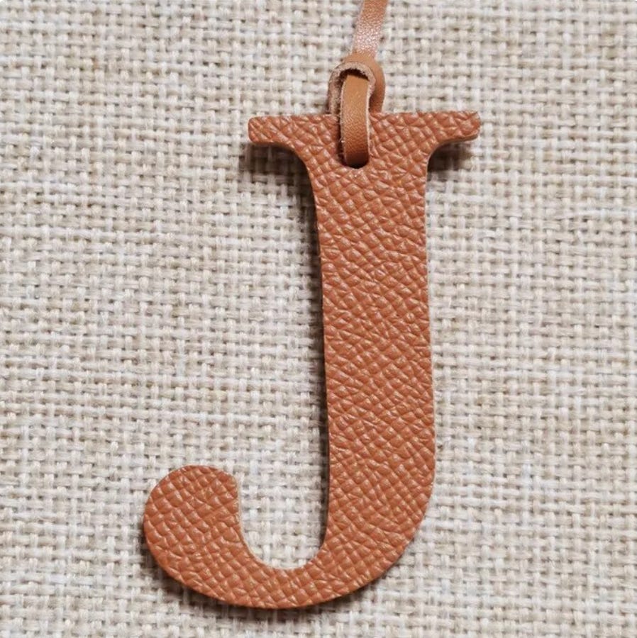 The letter J cut out of light brown leather with a looped cord of leather coming out of the top