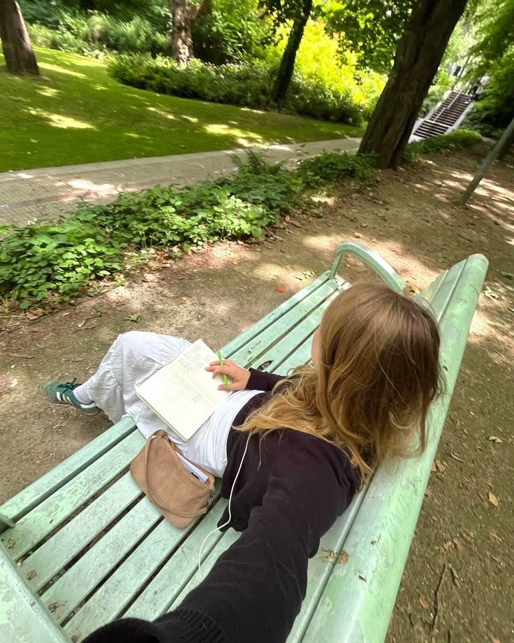 This may contain: a woman sitting on top of a green bench next to a lush green park filled with trees