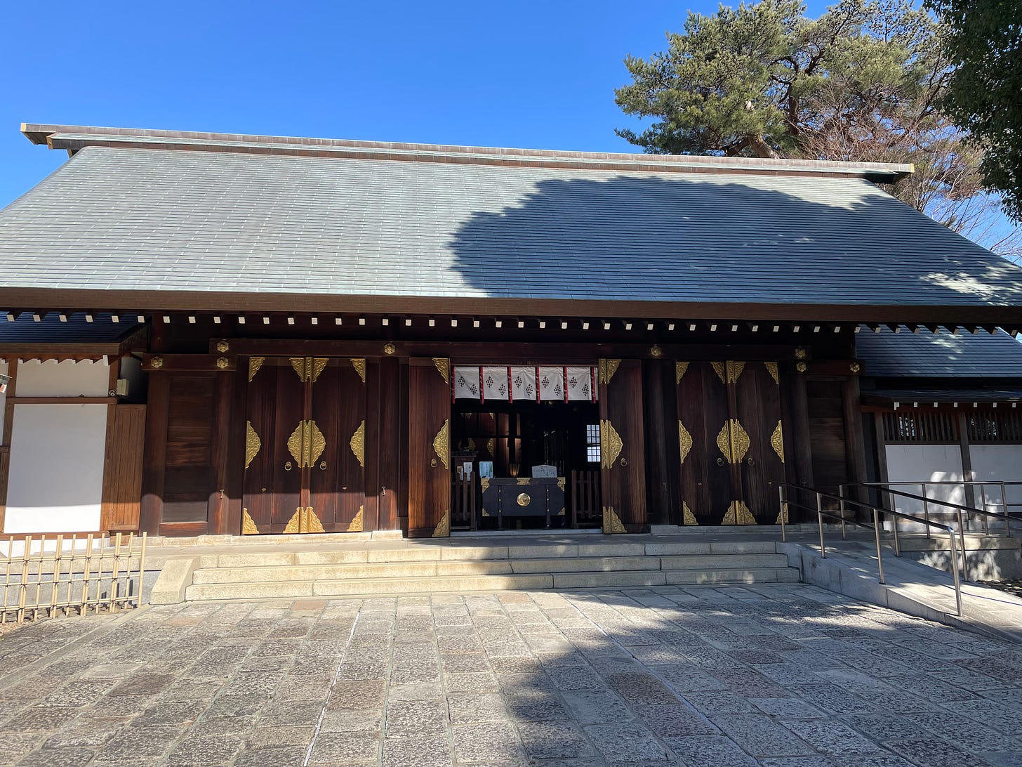 Main building of Shoin Shrine. Dark wood panels with golden trim on the outside. A sweeping roof. A red, white, and black noren curtain hangs over the door that houses the offering box and inner shrine elements.