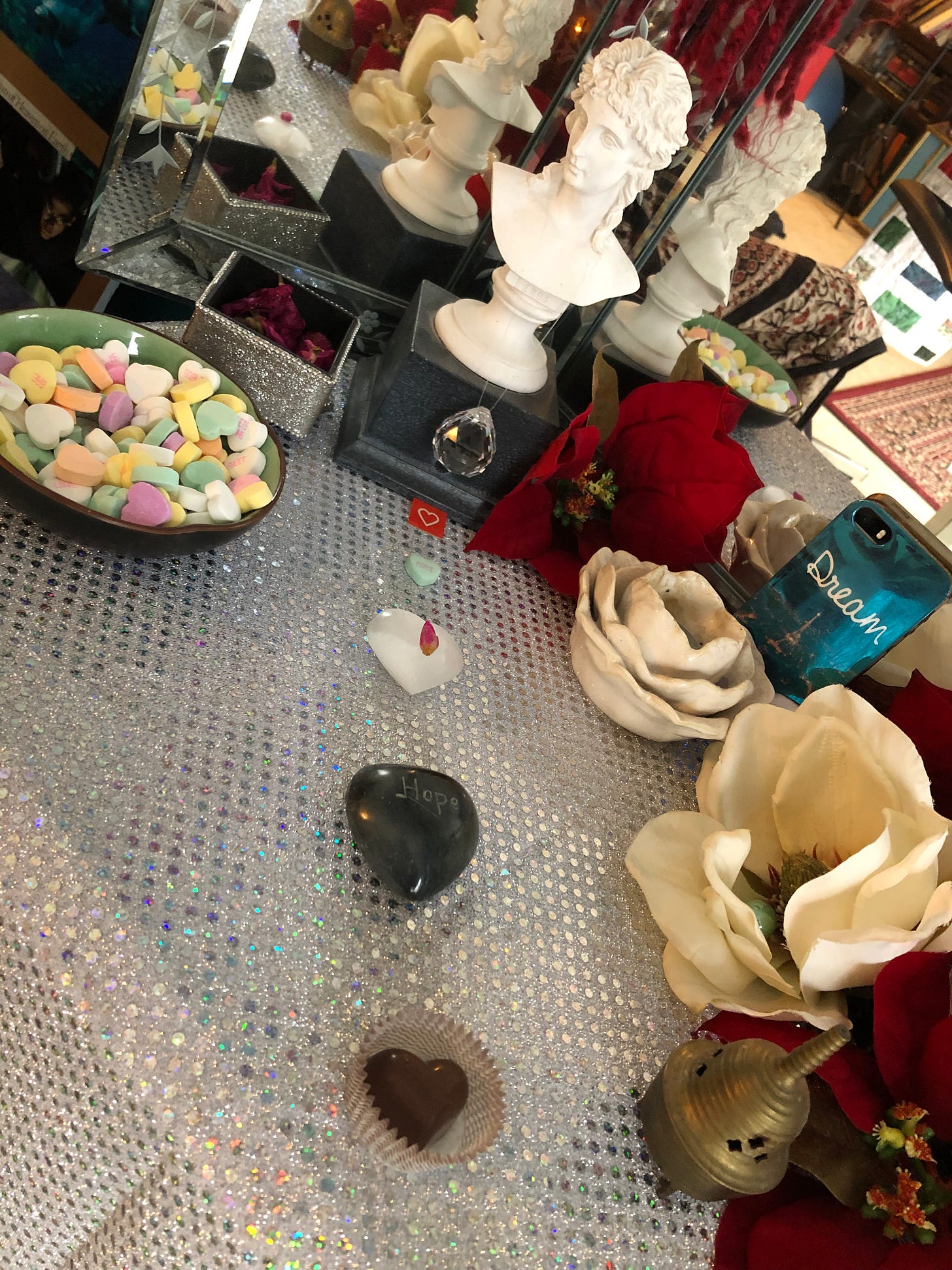 A bust of Eros, God of Love, overlooks an altar filled with silk flowers, crystals, and a bowl of candy conversation hearts. A line of other hearts from the chocolate to the stone leads from where you sit up to Eros. One pink flower bud dots a white crystal heart. A shiny teal phone case says, "Dream." And a polished gray stone heart says, "Hope."