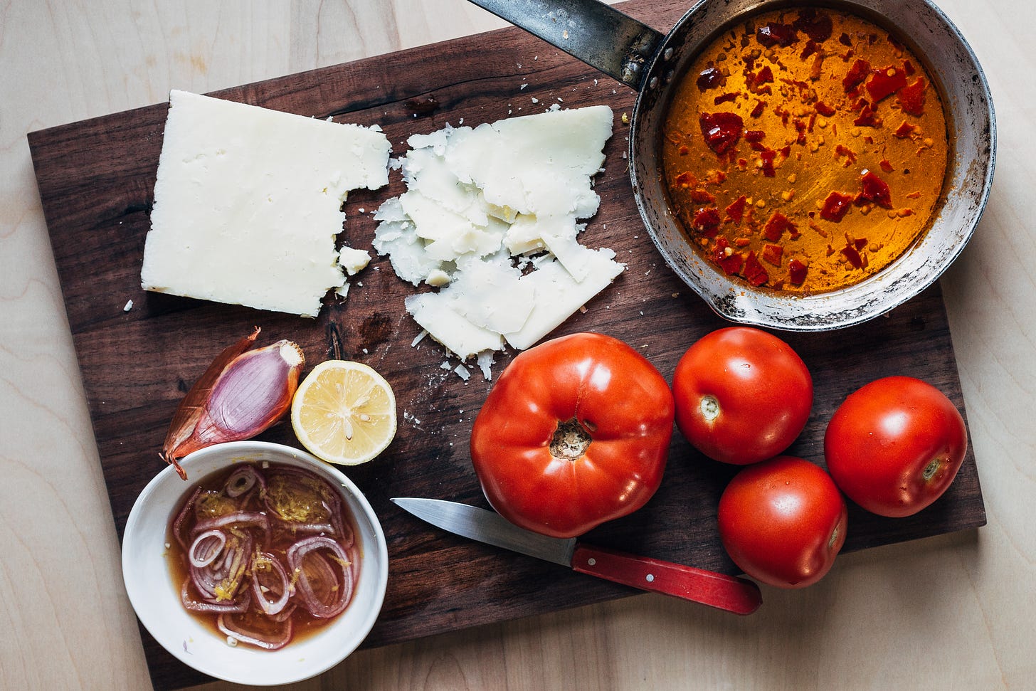 A cutting board with all the salad ingredients – olive oil in a skillet, whole tomatoes, shaved cheese, and shallots in a little bowl. 