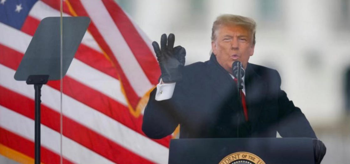 Donald Trump on January 6 2021, wearing black gloves, standing in front of the American flag.