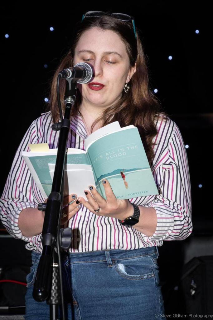 Me reading at the Coppenhall Open Mic in a striped shirt, and blue jeans, with my glasses on my head and red lipstick. Holding a copy of my poetry book 'It's All In The Blood'.