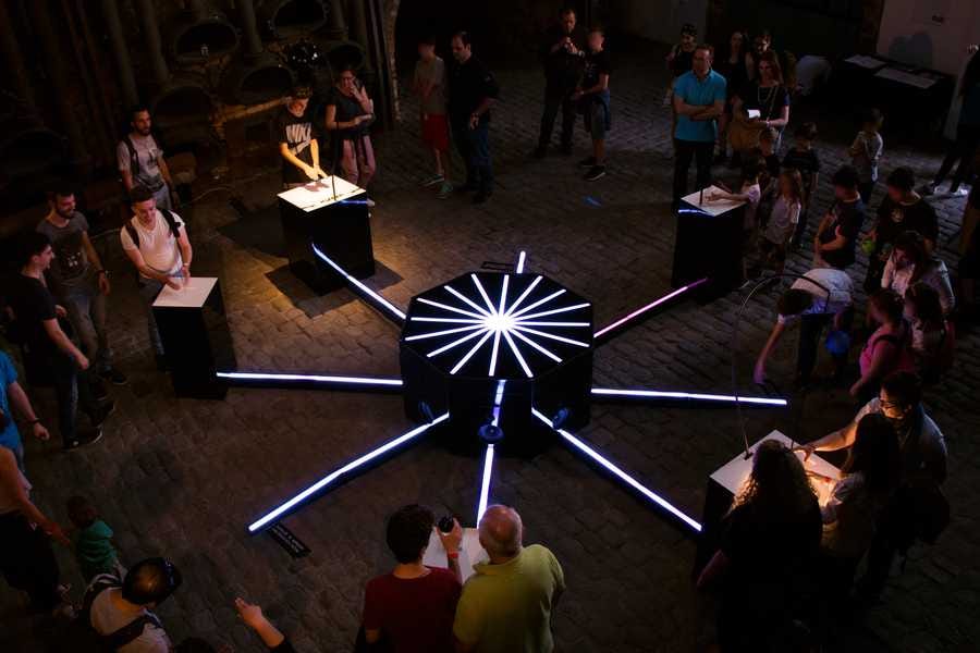 Cave of Sounds exhibited at Athens Science Festival 2018. An octagonal interactive installation is surrounded by many people in various stages of engagement. Photo: Anastasia Alekseeva.