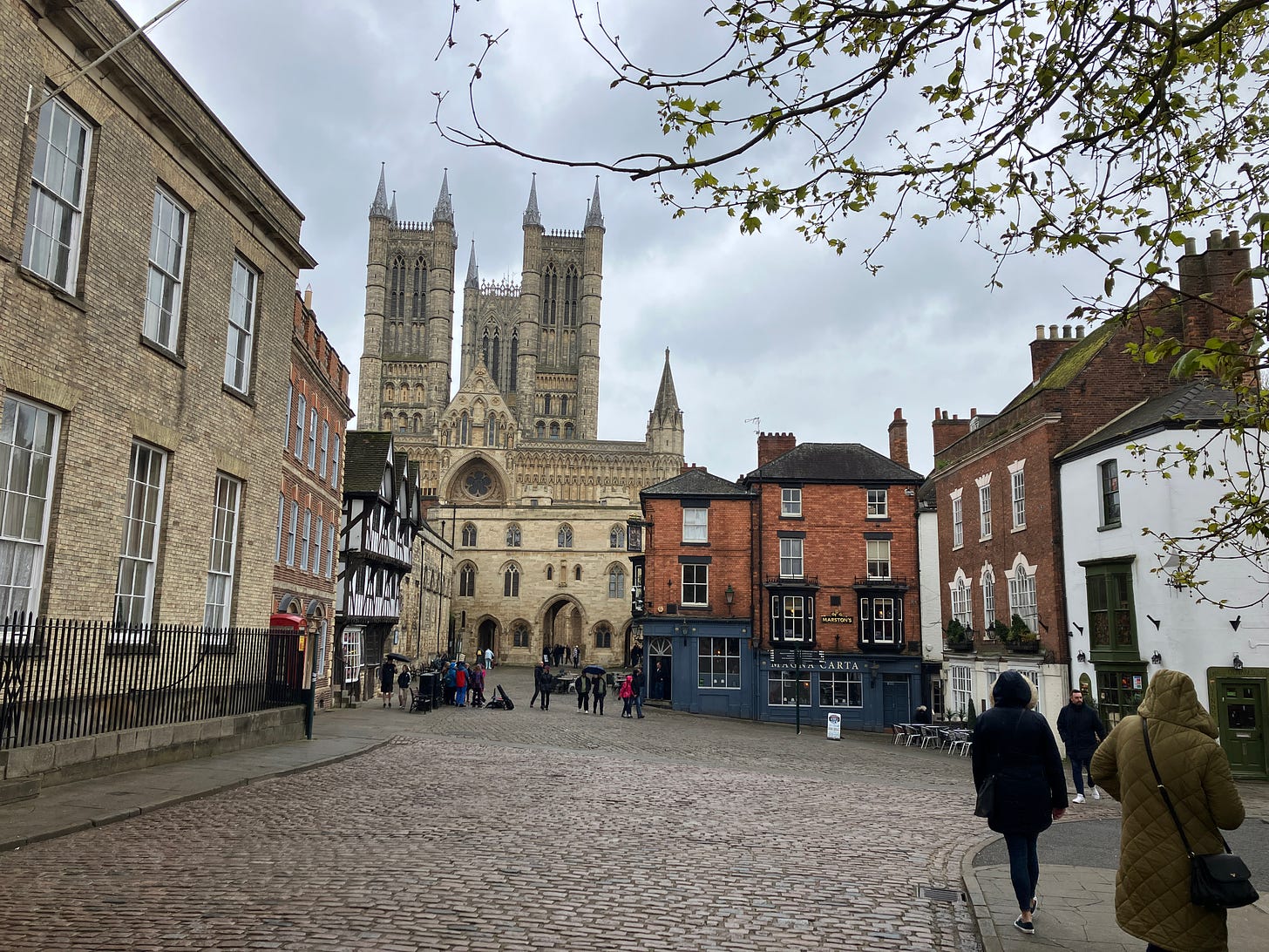 Lincoln Cathedral towers over surrounding historic buildings, with people walking on a cobbled street on an overcast day.