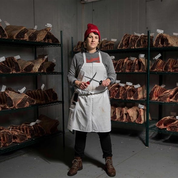 Image of a woman standing in front of racks of meat cuts, wearing a white apron and a read hat, and holding a knife with a sharpening implement.