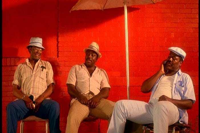 This image shows Do the Right Thing's three Corner Men sitting on chairs in front of a fire-engine-red wall and underneath an umbrella. Left to Right: M.L. (played by Paul Benjamin), Sweet Dick Willie (played by Robin Harris), & Coconut Sid (played by Frankie Faison). Coconut Sid holds his right hand to his right cheek, wiping perspiration from his face.