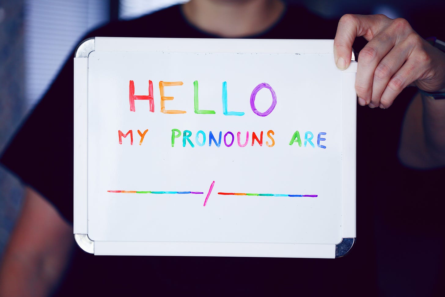 Person holding a white board with the following text written in rainbow colors: "Hello my pronouns are __/__."