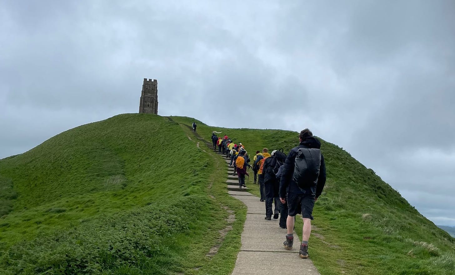 Walkers in single file, walking on a concrete path up a green hill with a church tower on the top