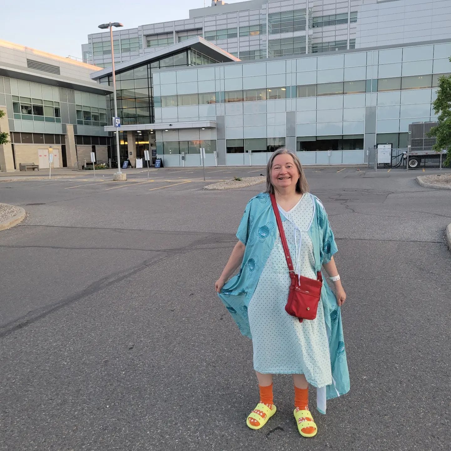 woman wearing a hospital gown and another gown as jacket, with yellow crocs and a red purse over the shoulder. She stands in front of the Ottawa General parking lot and smiles