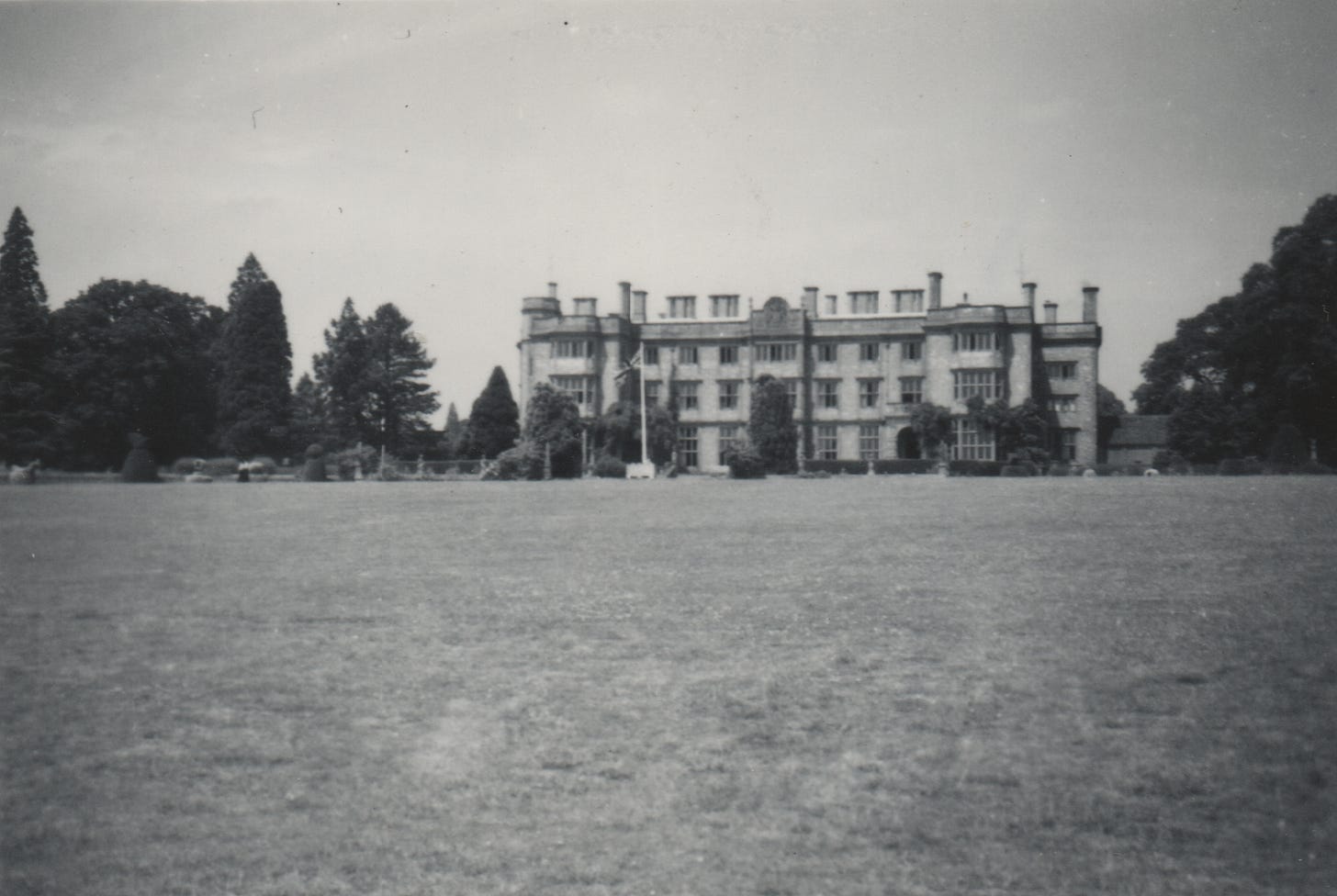 A large stately building with a landscaped topiary either side. In the foreground is a large lawn. 