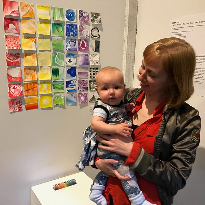 A seven by seven grid of monochromatic paintings, or One Tiny Painting a Day Seven Days a Week for Seven Weeks by ilyse iris, who is also pictured holding an infant.