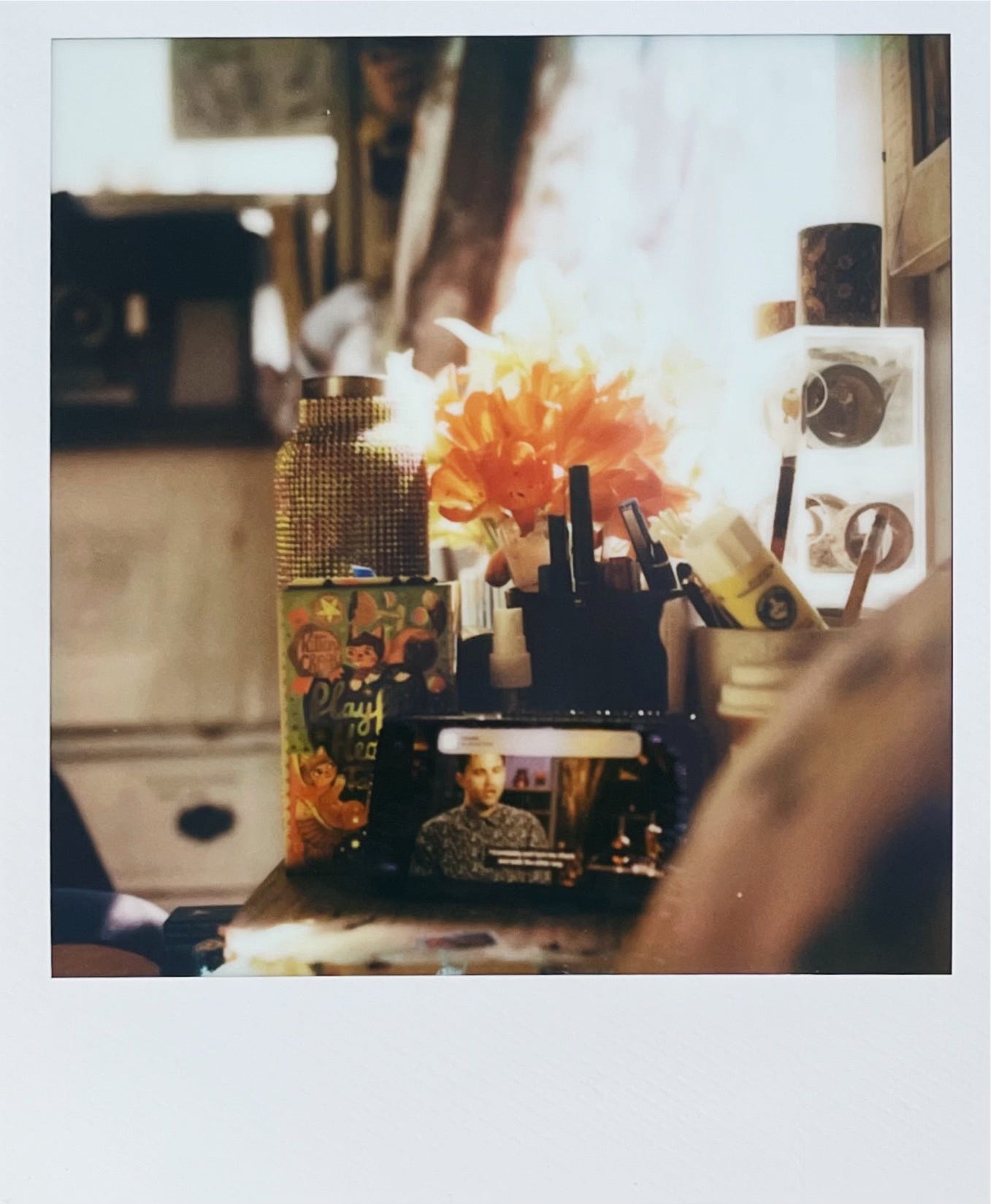 A cozy, cluttered desk with orange flowers, art supplies, and a phone streaming a man speaking in an interview.