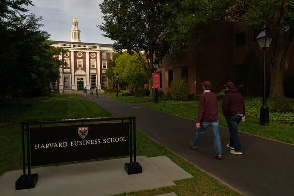 Two people wearing maroon tops and blue jeans walk along a path in the foreground, near a sign for Harvard Business School. A building with red brick and white trim stands in the background.