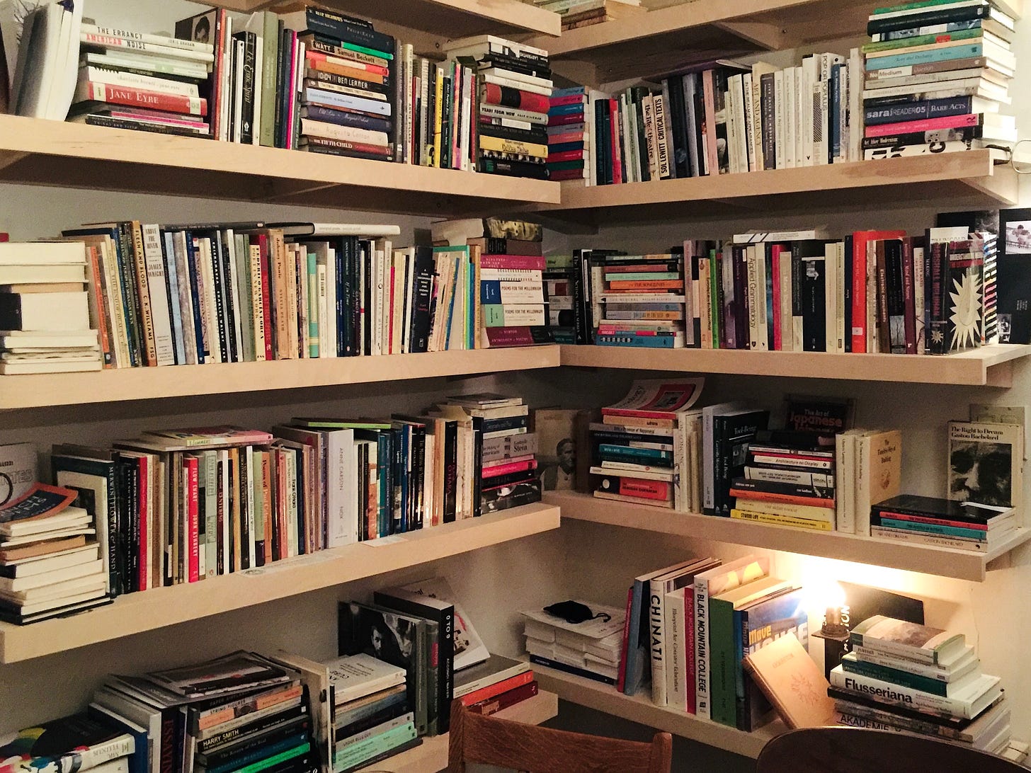 Photograph of the corner of an idiosyncratic library. The pale wood shelves hold books in stacks and upright. You can make out the titles of a few of the many books including Jane Eyre, two volumes of Poems for the Next Millennium, Nox, The Making of Americans, Chinati, Black Mountain College, and Flusseriana. 