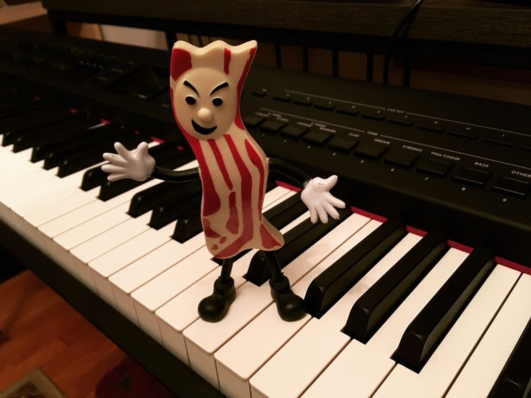 An anthropomorphic strip of bacon with a face, arms, cartoon gloved hands and shoes standing atop a piano keyboard in all its glory.
